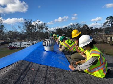 Volunteers in safety gear placing a blue tarp on a roof
